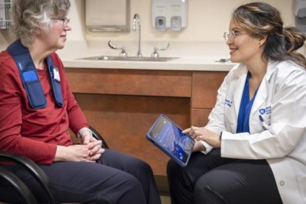 A physician sits next to a patient in a clinic room. The physician holds a tablet computer as she smiles and looks at the patient.