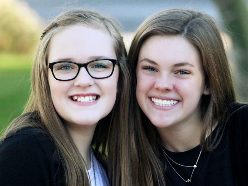 Two adolescent girls with light hair sit close together a smile outdoors on a bench.