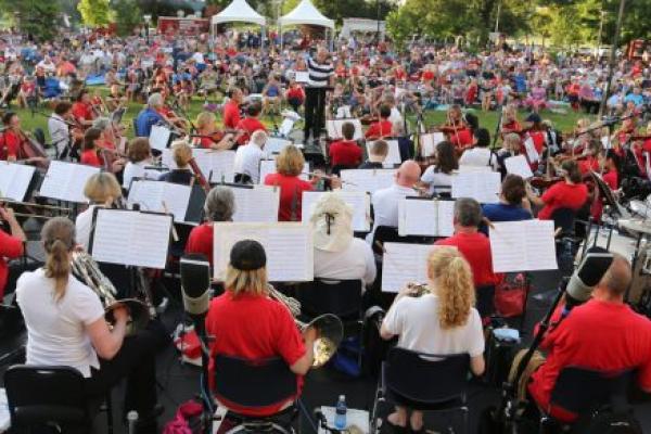 Penn State Health Milton S. Hershey Medical Center is lit up in red and white lights as an orchestra plays on a lit stage in front of the building's signature Crescent. A crowd of a couple thousand people looks on.
