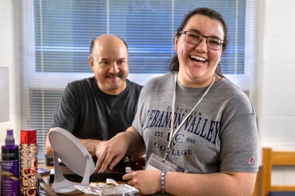 A young woman laughs while arranging items on the top of her dresser. She is wearing glasses and a name badge lanyard around her neck. A man stands behind her, watching and smiling.