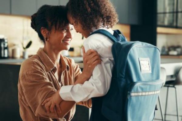 Mom and girl touch foreheads as girl gets ready for school. Girl is wearing a school uniform and backpack.