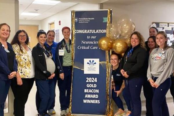 Thirteen members of the Neuroscience Critical Care team at Milton S. Hershey Medical Center smile as they stand around a pull-up banner that reads “Congratulations to our Neuroscience Critical Care – 2024 Gold Beacon Award Winners.”