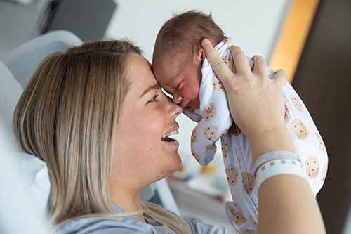 Mother smiles at baby as she holds him up so their foreheads and noses touch. The mother sits in a hospital bed.