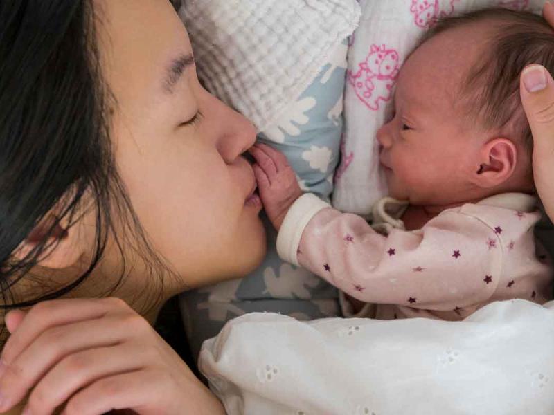 Young mother adoring her newborn baby. Her hand is gently resting on the baby’s head as they are both lying in bed close to one another.