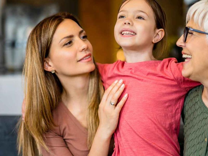 A smiling grandmother is embracing her daughter and granddaughter in a gentle hug while relaxing at home.