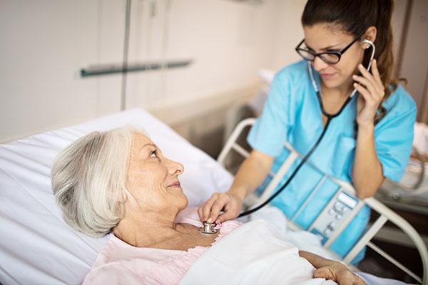Female nurse examining senior woman lying on bed. Medical worker is  with stethoscope in hospital. Elderly woman is looking at professional.