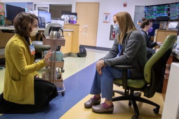 A woman in a cardigan sweater kneels in a hospital common room. She speaks with a woman in scrubs and a sweater sitting in a chair in front of her. Next to the kneeling woman is a rack of coffee pots and other implements. Behind the seated woman in a bank of computer screens, and another woman sits at a desk. Everyone is masked.