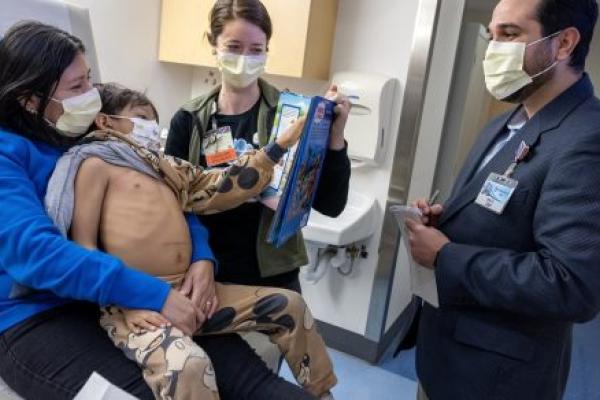 Four people in a hospital room talk behind surgical masks. One is a child, who sits on a woman's lap. The child's shirt has been pull aside. He gestures at a man who stands taking notes on a pad.