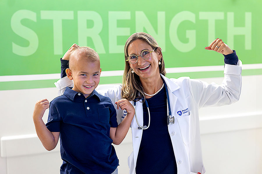 A woman in a white lab coat and a boy stand beside a green wall with the words strength displayed on the wall, smiling and engaged in a discussion.