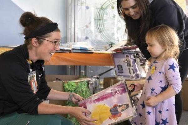 A woman kneels down to hand a packaged princess costume to a young girl while another woman leans over as she looks on.