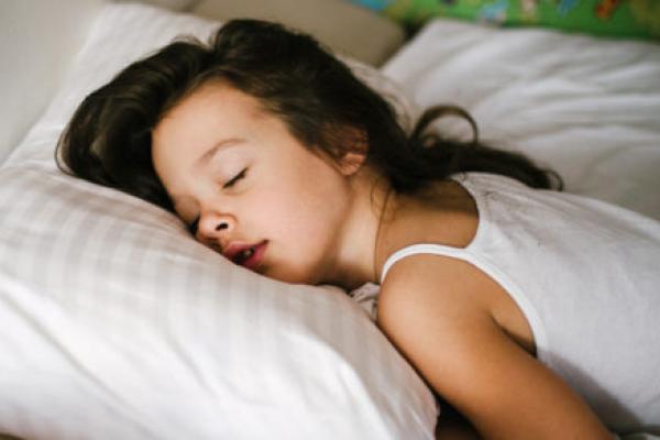 A young girl sleeps in bed, with her head on a pillow. Her head is facing the camera.