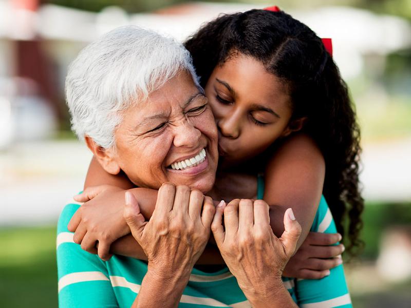 A loving teen girl embracing and kissing her happy grandmother from behind and holding each other outdoors.
