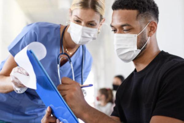 A male patient signs a document on a clipboard which is being held by a female medical staff member wearing scrubs and a stethoscope.