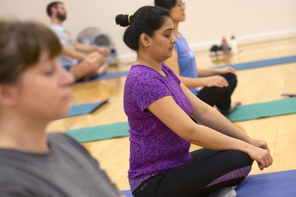 A group of individuals seated on yoga mats in a bright room, practicing meditation or mindfulness exercises, with a focus on a woman in a purple shirt in the center.]
