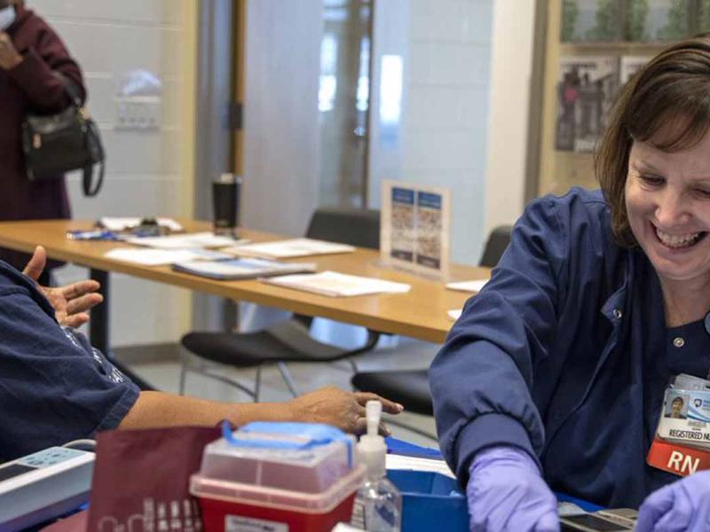 Penn State Health Community Health Nurse prepares to provide a vaccination to a community member. They are sitting at a table covered in medical supplies and are talking and smiling.
