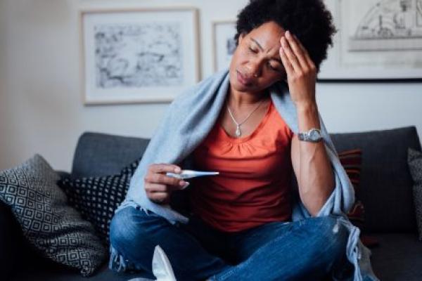 A young woman seated on a couch clutches her forehead and reads a thermometer. A box of tissues is positioned near her lap and a blanket is draped over her shoulders.