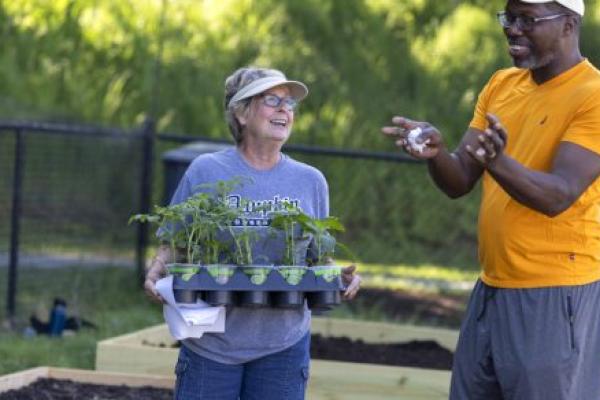 A man and a woman stand side-by-side talking in a community garden. The woman is holding a container with several potted plants. Both individuals are smiling.