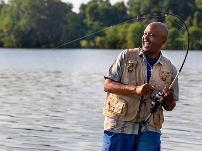 Heart transplant recipient, Dave, dressed in a fishing vest, stands by a lake and smiles as he reels in his fishing line. His fishing rod is bent over his head.