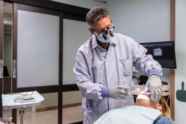 A physician with a tooth extraction tool tends to a patient who is reclined in an exam chair. A tray of tools and a computer screen with an X-ray are in the background.