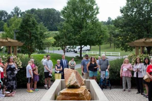 Approximately 20 people stand on a brick patio, surrounding a stone memorial, at a ceremony.