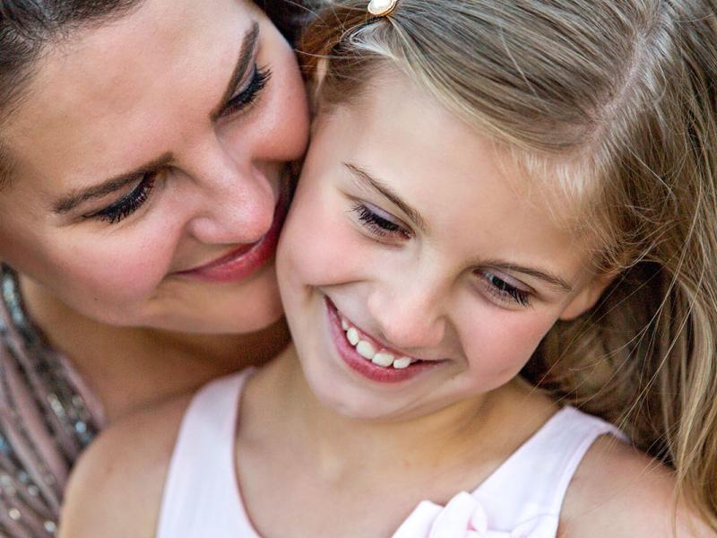 A girl and her mother are outside in a field. The mother is whispering something into the girl's ear.