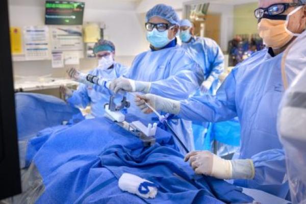 A team of medical professionals in full surgical attire, including masks, gowns, and protective eyewear, performs a minimally invasive procedure in a hospital operating room.