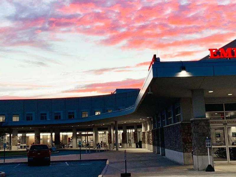 A hospital’s interior is illuminated as lights come on in the building at dusk. Clouds dot the sky. A bell tower rises above the hospital on the left. On the right, an emergency sign marks the entrance below the hospital logo. A circular garden with shrubs and a sign sits to the left of a parked truck.