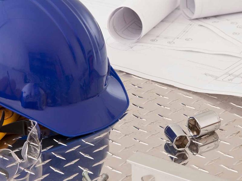 A variety of construction work tools positioned on the surface of a tabletop workstation, including construction blueprints, a hardhat, work gloves, screwdriver and a tape measure.