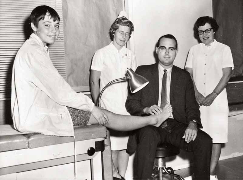 Dr. Willis Willard touches the foot of a boy who is seated on an exam table. Two nurses wearing short-sleeved uniforms stand on either side of Dr. Willard. He is wearing a suit, tie and glasses. The boy is smiling and wearing a jacket with a string hanging down. The nurse on the left is wearing a nurse’s cap and has her eyes closed. The nurse on the right is wearing horn-rimmed glasses. 