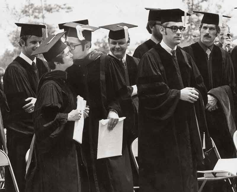 Eight medical students stand and wear caps and gowns at Penn State College of Medicine’s first graduation ceremony in 1971. Folding chairs are behind each of them. On the left, a man kisses a woman on the cheek.