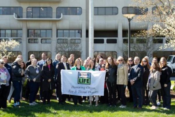 A group of people pose with a “Donate Life” flag. A hospital building with windows is in the background.