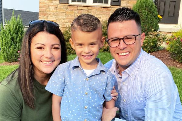 A woman and man kneel on either side of a young boy in the front yard of a house. The woman wears a t-shirt and jeans with a hole in the knee. She has sunglasses on her head. The man wears a long-sleeved shirt, pants and glasses. The boy wears a short-sleeved shirt and shorts.
