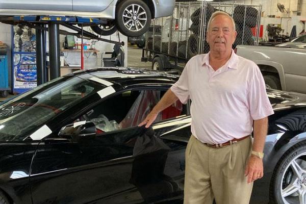 In a commercial garage, a man stands in front of a car with his hand on the open window. In the background on the left, a car sits atop a riser. Behind it is an office with a desk and chair. On the right, several vehicles surround a gated enclosure filled with tires.