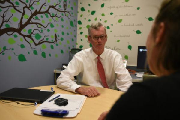 Dr. George Blackall, PsyD, MBA, is seen sitting at table counseling a female patient. He is looking directly at her. The patient Is facing Dr. Blackall and her face is not visible. The room has a decal of a tree on its walls.