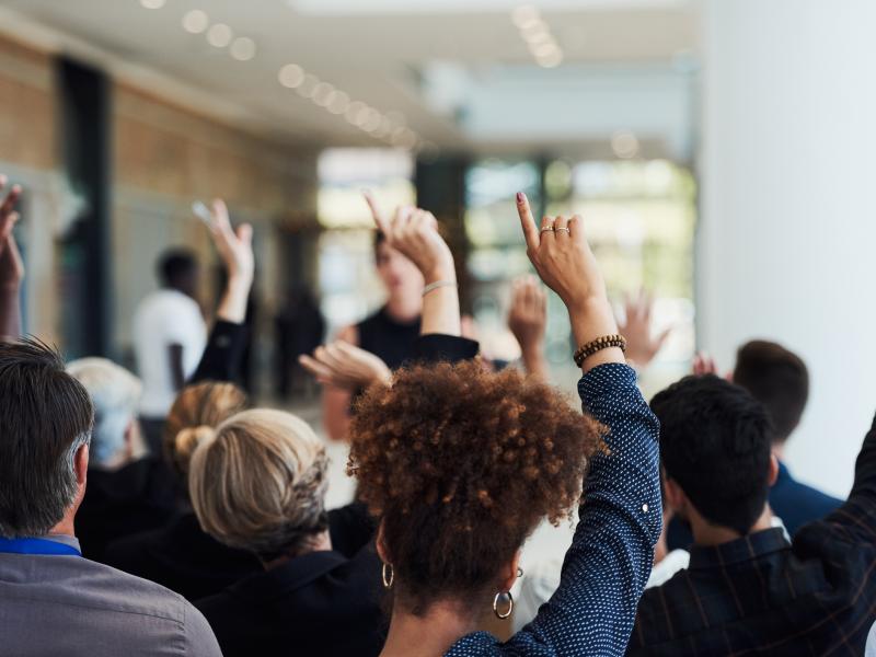 An audience with its back to the camera raises their hands to ask a question.
