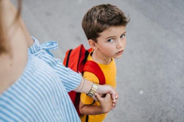 A boy wearing a backpack looks up at mother sadly as he holds her hand.