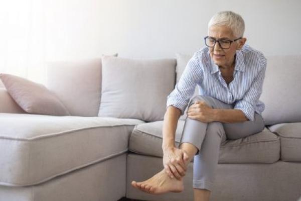 An older woman suffering from leg pain holds her ankle at home.