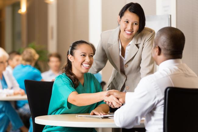Two physicians sitting at a table shake hands while another who's standing looks on.