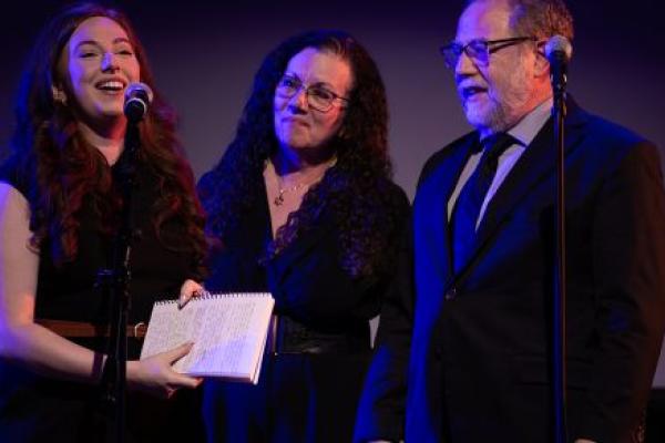 Two ladies and one gentleman stand side-by-side. One lady is speaking into a microphone and smiling. The other lady and the gentleman hold hands as they look at the one speaking.
