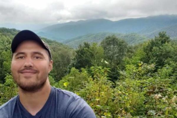 A young person is standing outdoors in front of foliage with a view of mountains and clouds in the distance.