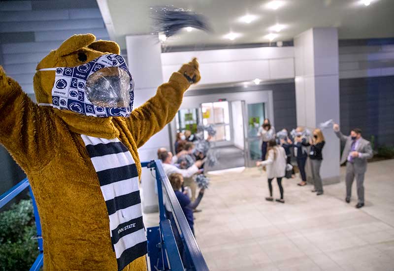 The Nittany Lion mascot, wearing a Penn State face mask, raises his arms. Below him, in an out-of-focus shot, hospital leaders wave pom-pom shakers to greet employees as they enter the new Penn State Health Hampden Medical Center on opening day, Oct. 1, 2021.