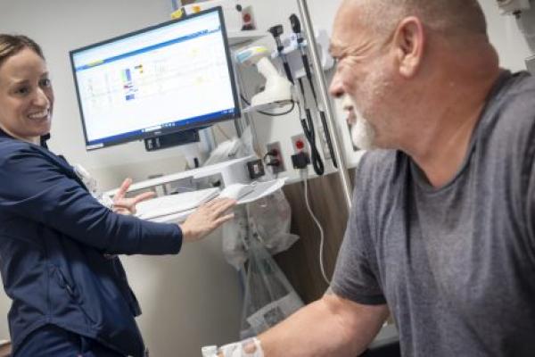 A nurse stands at a computer, looking to the side as she speaks with a patient. They are in a hospital emergency room treatment bay.