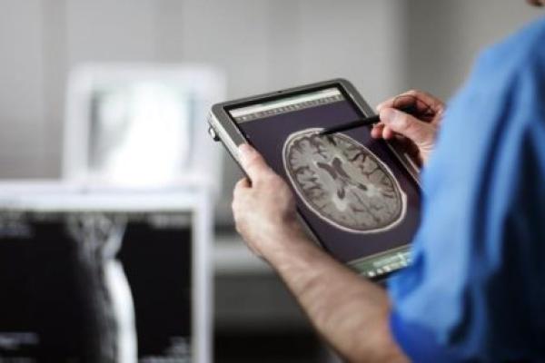 A healthcare professional in blue scrubs examines a brain scan on a digital tablet using a stylus.