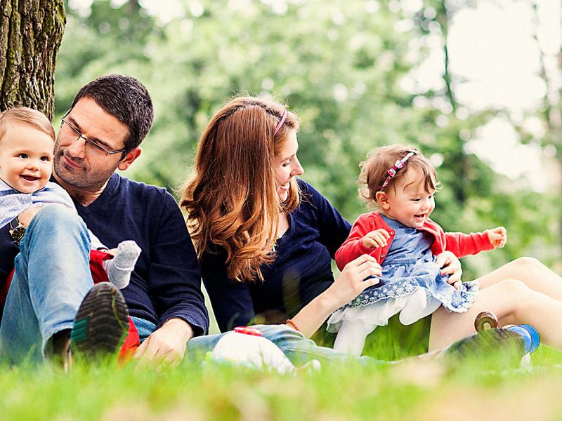 A man and woman each hold a small child on their lap as they sit in the grass next to a tree. Other trees are visible in the background.