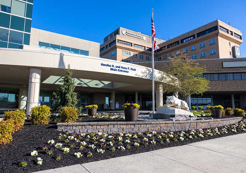 Exterior of Penn State Health Holy Spirit Medical Center. The main entrance sign reads “Hershey H. and Hasu P. Shah Main Entrance.” A Nittany Lion statue is next to a flagpole with the American flag. A flowerbed is in the foreground. The Penn State Health Holy Spirit Medical Center sign is on the top of a brick section of the hospital.