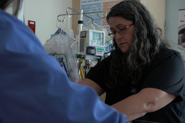 a female respiratory therapist wearing glass and navy scrubs gathers supplies in a hospital room. Medical monitors are in the background. A hospital employee in a blue shirt is in the foreground.