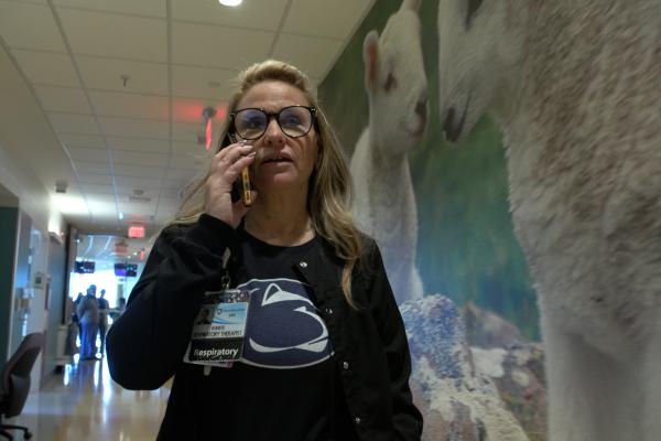 A female respiratory therapist wearing a navy blue Penn State shirt walks down a hospital hallway while talking on her cellphone. A large mural with lambs on it is in the background.
