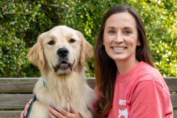 A golden retriever dog named Thor is on the left and a women wearing a pink top is on the right. They are sitting on a wooden bench with trees in the background. Both are looking at the camera. This photo was taken in August 2023.