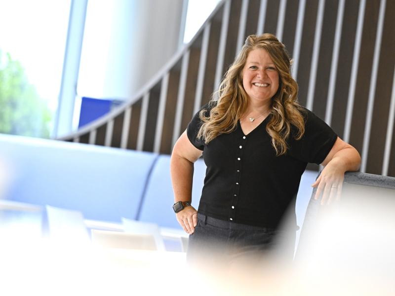 Katy Rush, a senior recruiter for Penn State Health, stands in front of seats in the lobby of a health system building. She is smiling and wearing a button-down blouse and pants. She has one arm one on the seat and the other by her side. Trees are visible through a window on the left. 