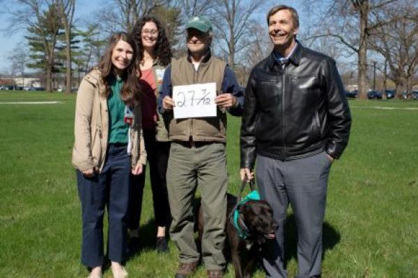 Two women and two men smile as they pose for a photo outdoors on the Milton S. Hershey Medical Center campus. The man in the middle is holding a sign that reads 27-7/12. The man on the right is holding a service dog’s collar.